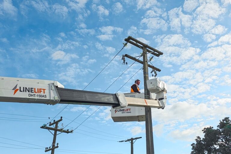 An upward angle of a Linelift® elevated work platform with its boom extended bucket positioned near a power pole against a blue sky