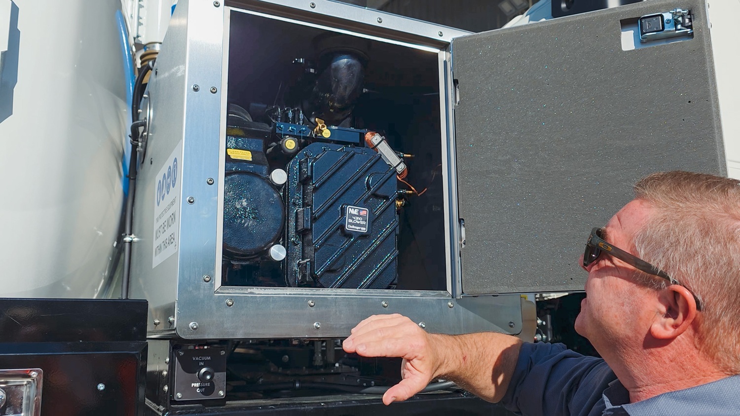 A man crouching next to a blue Moro Kaiser vacuum pump fitted on a vacuum tanker truck, talking to the camera.
