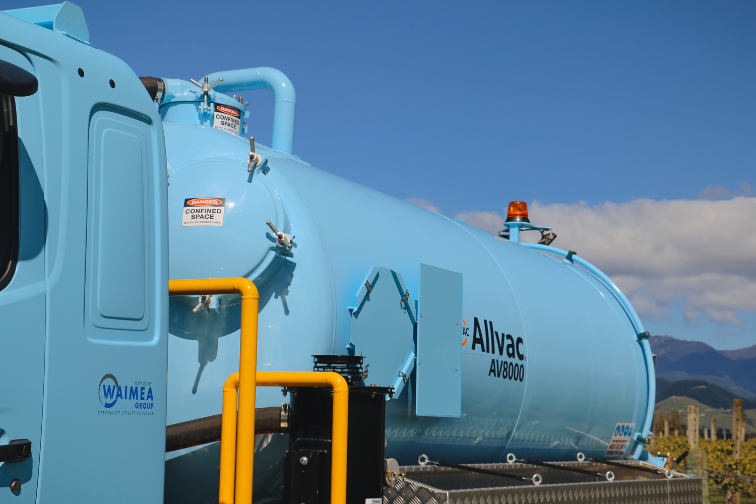 A vacuum tanker truck painted pale blue, with a large tank, side trays, and a compact chassis, parked in a green vineyard