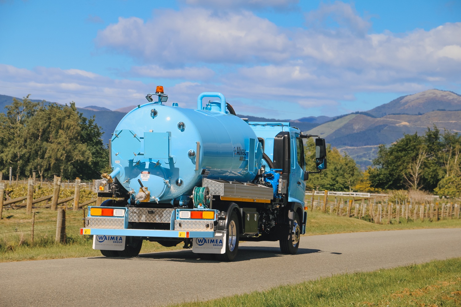 A vacuum tanker truck painted pale blue, with a large tank, side trays, and a compact chassis, parked in a green vineyard