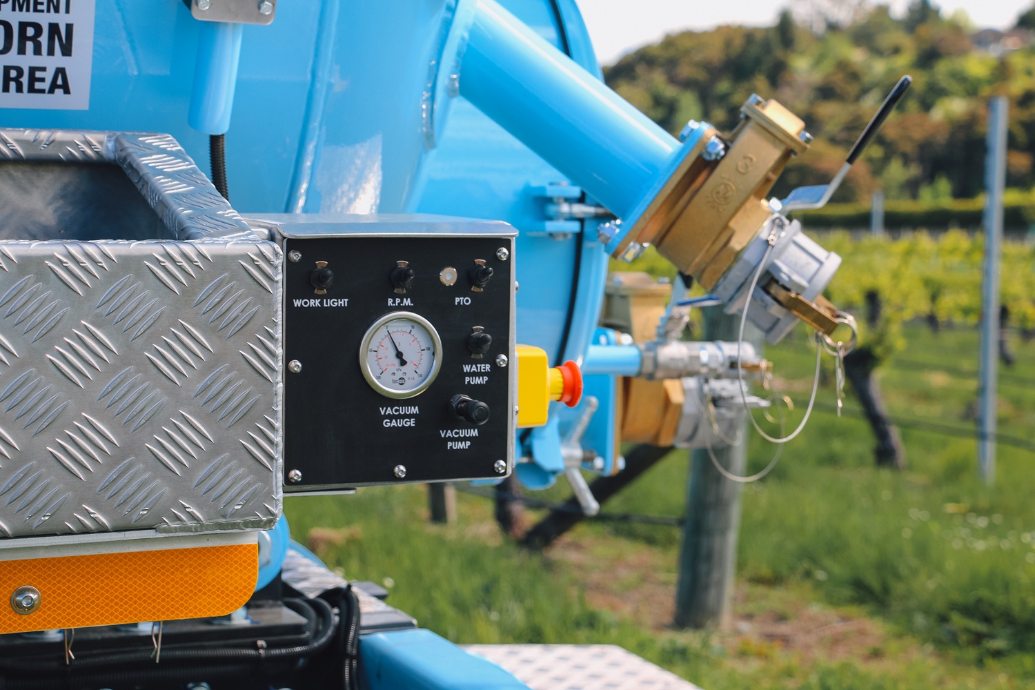 A vacuum tanker truck painted pale blue, with a large tank, side trays, and a compact chassis, parked in a green vineyard
