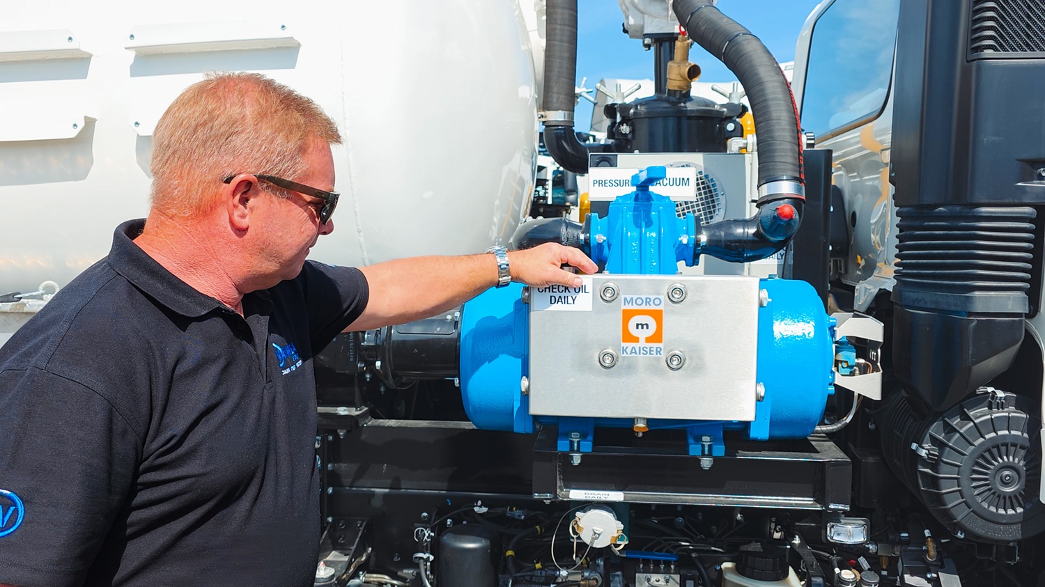 A man crouching next to a blue Moro Kaiser vacuum pump fitted on a vacuum tanker truck, talking to the camera.