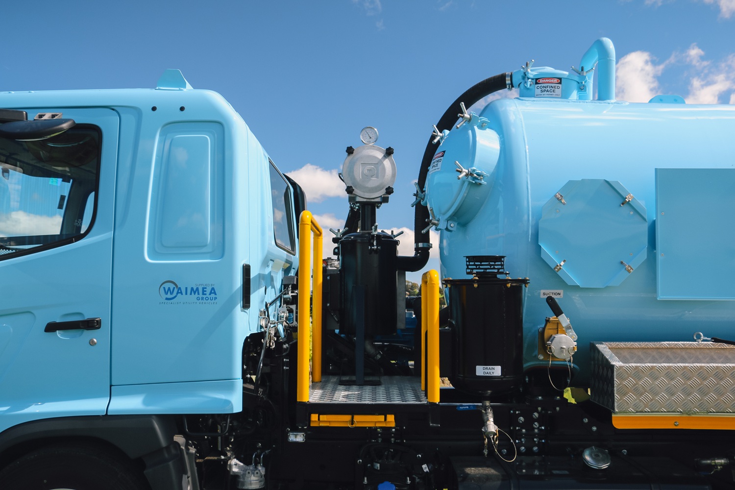 A vacuum tanker truck painted pale blue, with a large tank, side trays, and a compact chassis, parked in a green vineyard