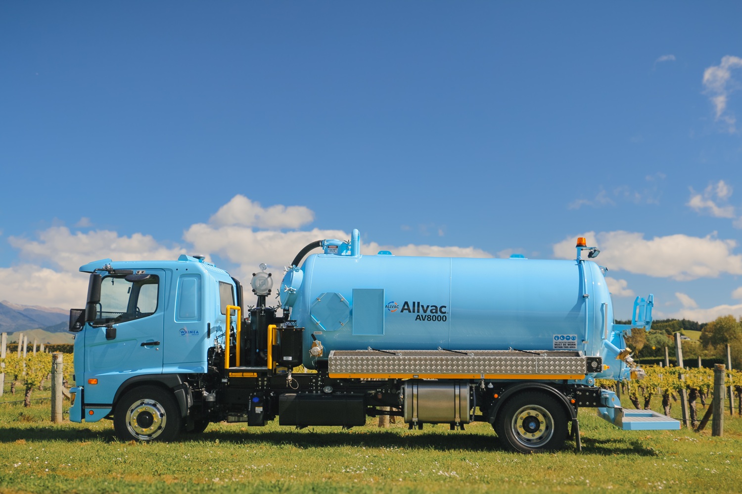 A vacuum tanker truck painted pale blue, with a large tank, side trays, and a compact chassis, parked in a green vineyard