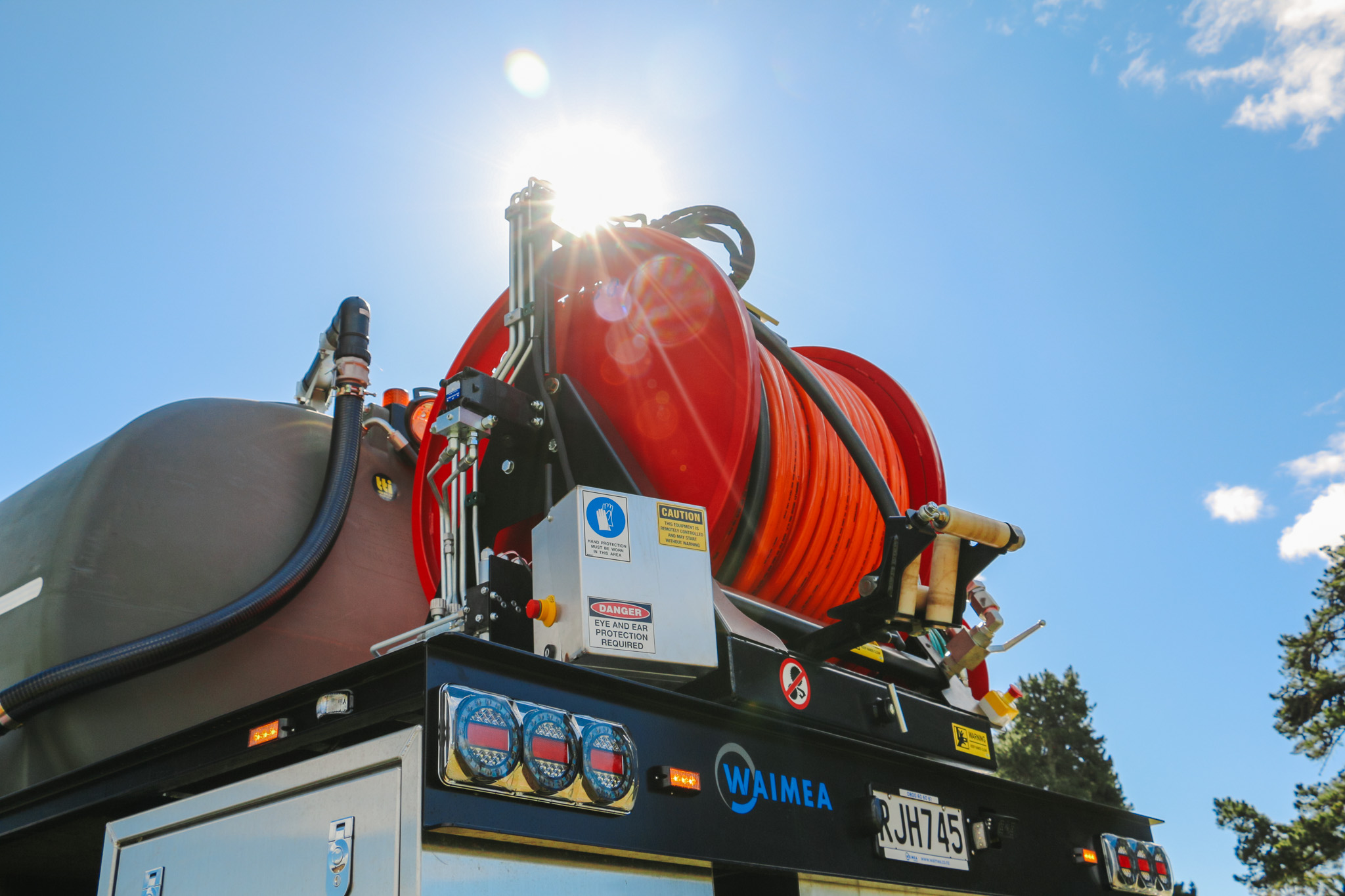 A large utility truck parked on the grass in front of a body of water, equipped with jetting equipment, carrying a 6000L tank and two hose reels with a set of nozzles for jetting out drains and culverts.