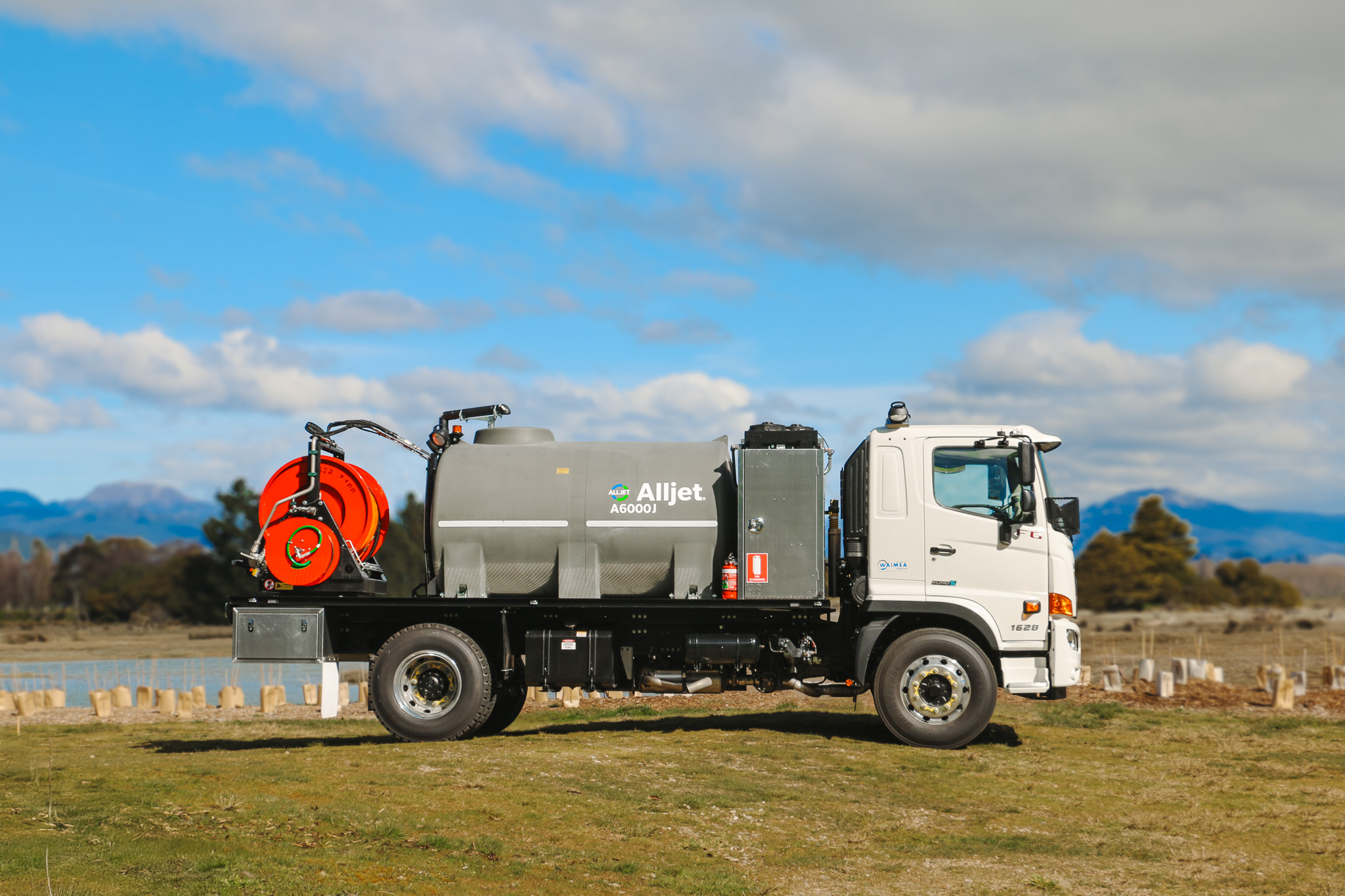 A large utility truck parked on the grass in front of a body of water, equipped with jetting equipment, carrying a 6000L tank and two hose reels with a set of nozzles for jetting out drains and culverts.