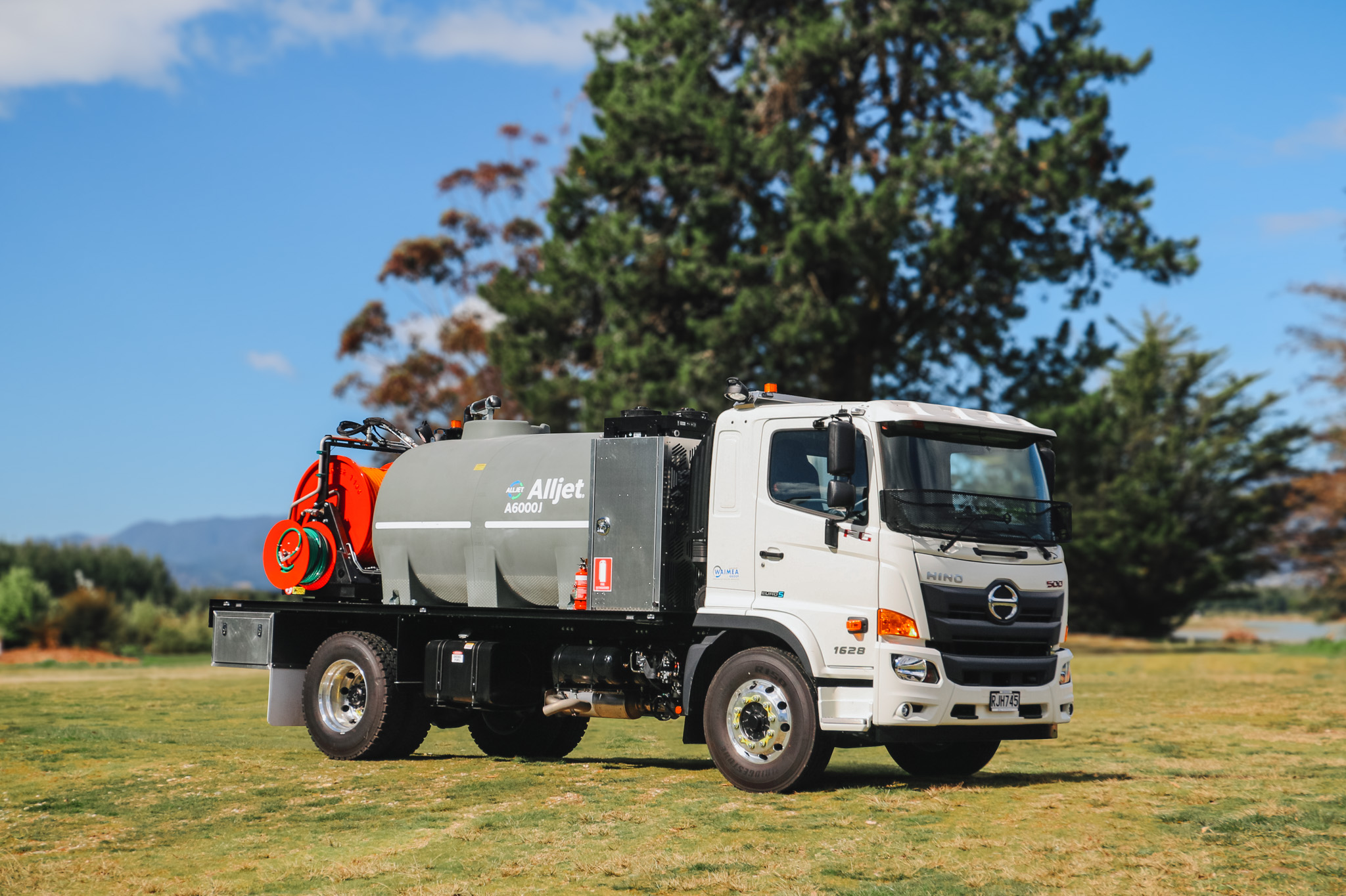 A large utility truck parked on the grass in front of a body of water, equipped with jetting equipment, carrying a 6000L tank and two hose reels with a set of nozzles for jetting out drains and culverts.