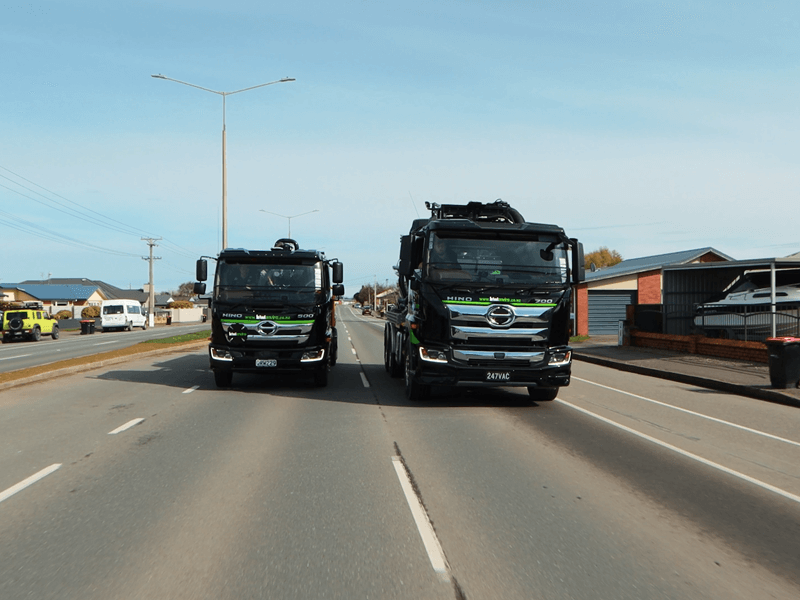 Two men, one wearing a hi-vis jacket with Waimea Group on the back, talking in front of an orange hydrovac being operated on a worksite.