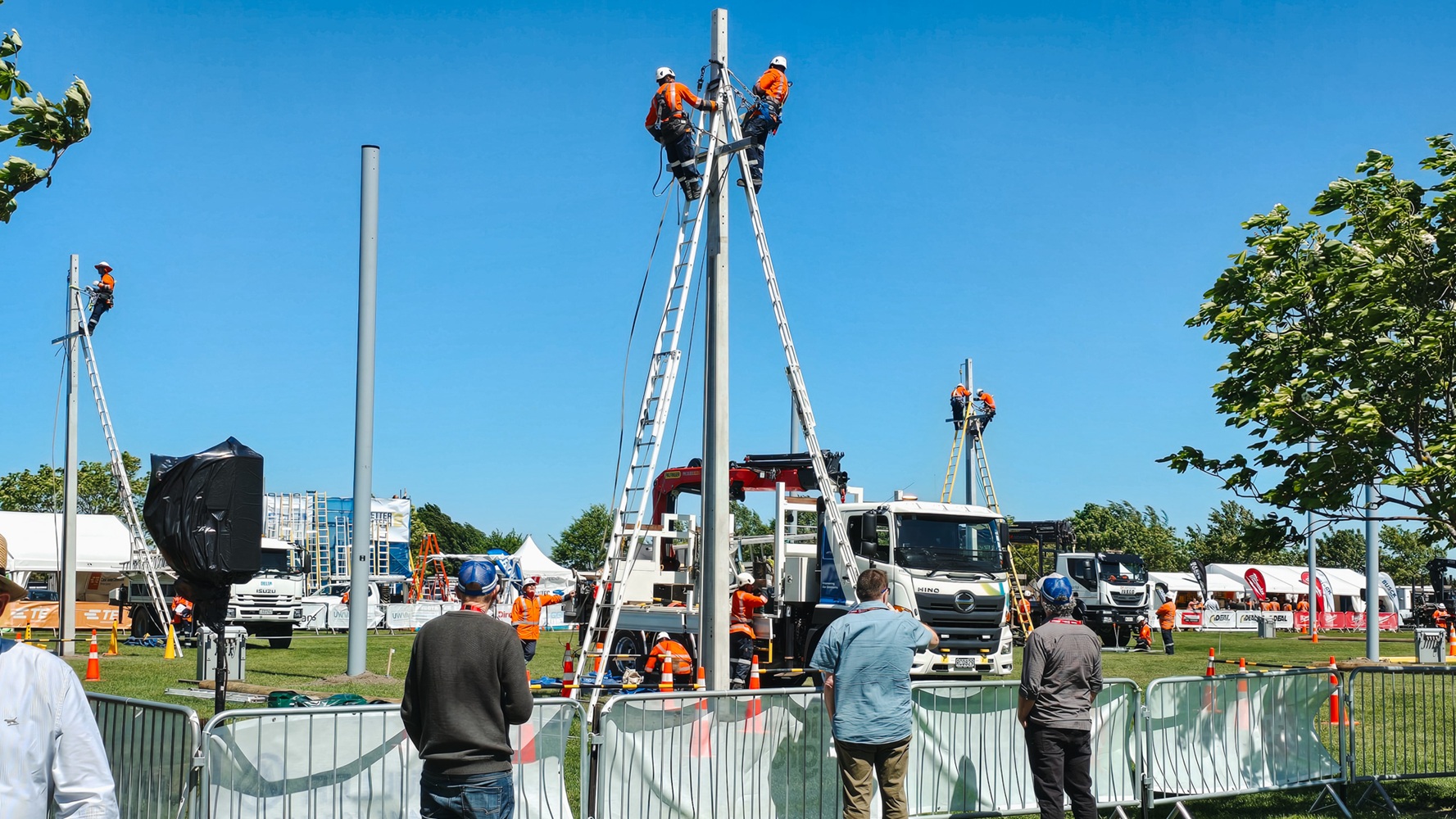 A showground viewed from up high, with lots of lines crane trucks set up with their crews demonstrating next to power poles, and stalls around the edge of the grounds, early in the morning.