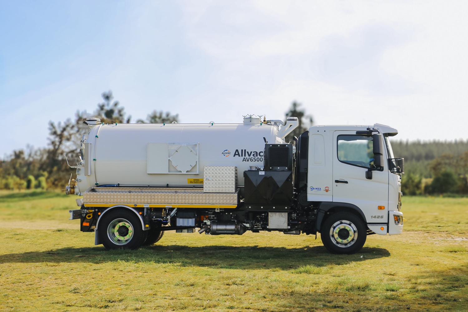 A large white vacuum tanker in the middle of a large field with trees and water in the background, with its tank tilted up on a hoist, facing diagonally away