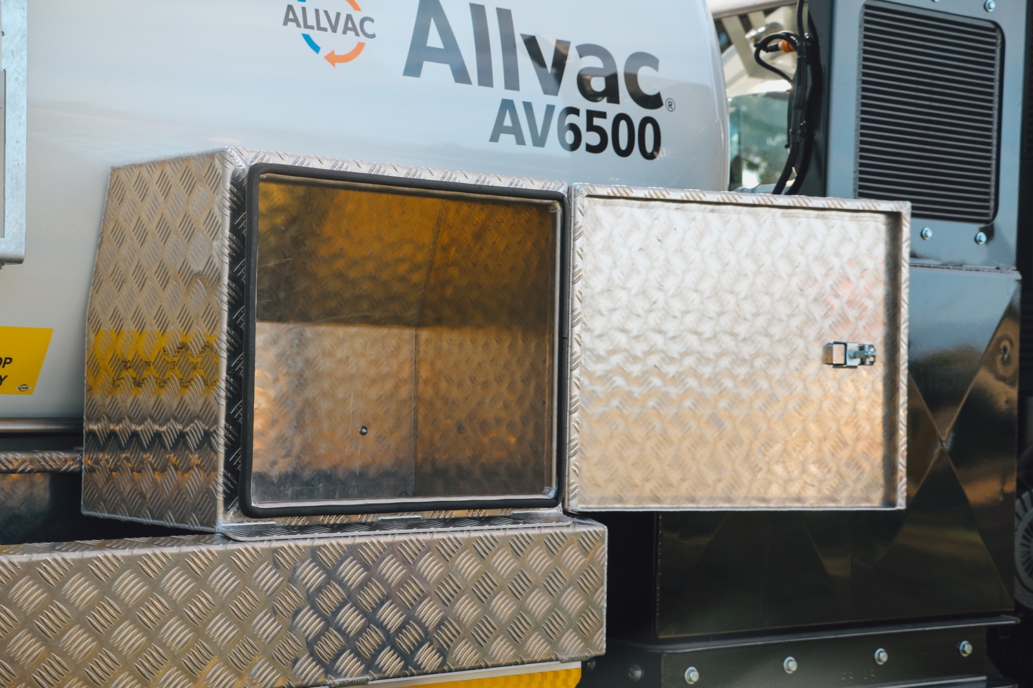 A large white vacuum tanker in the middle of a large field with trees and water in the background, with its tank tilted up on a hoist, facing diagonally away