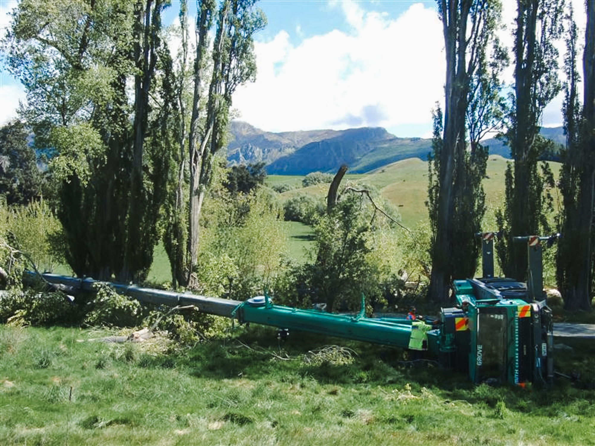 A specialist utility truck’s right rear outrigger is deployed on a grassy slope with a stabiliser pad and wedge to support it, with mudflaps on the wheels in the background with Waimea Group branding on them.