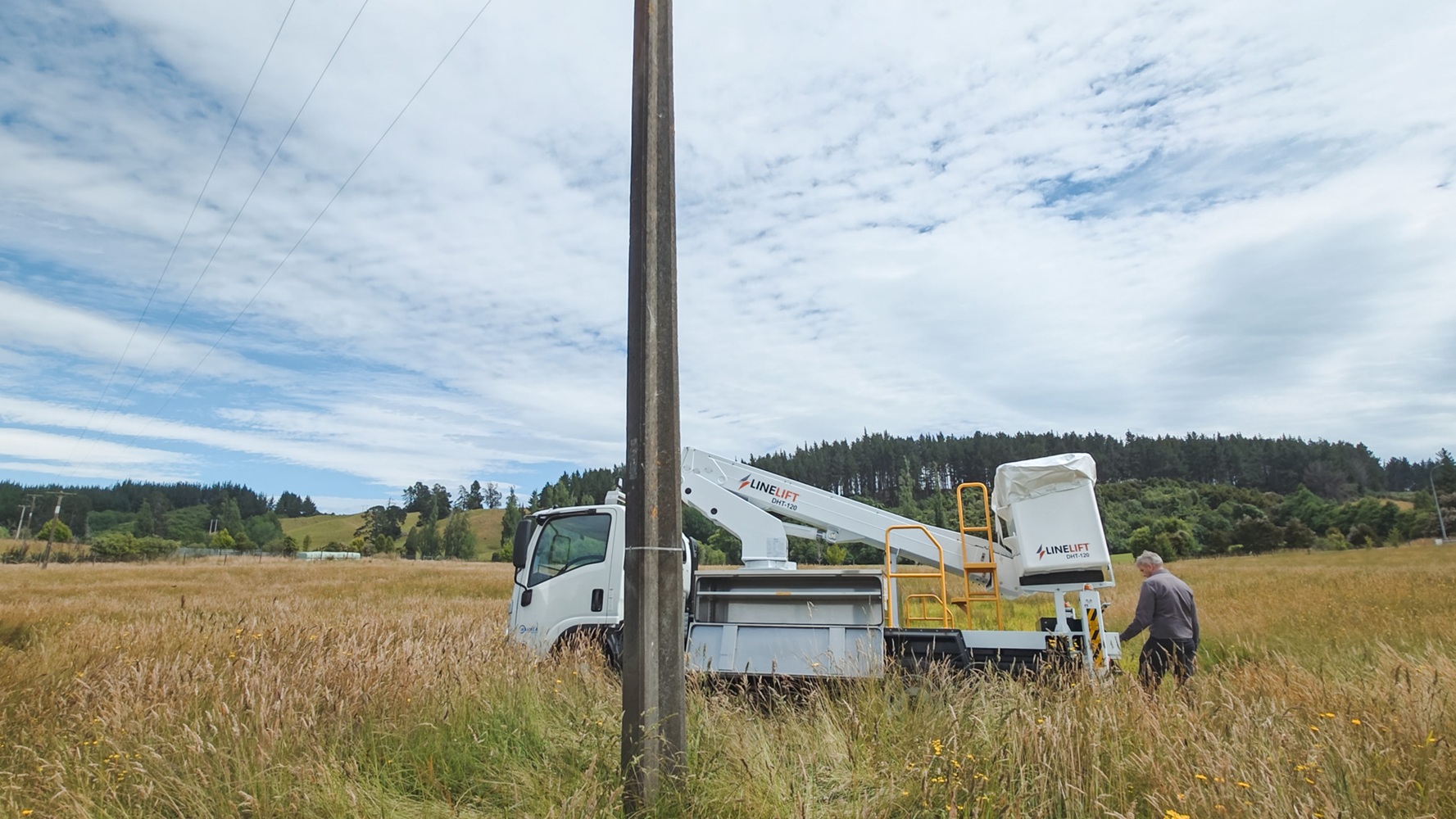 A specialist utility truck’s right rear outrigger is deployed on a grassy slope with a stabiliser pad and wedge to support it, with mudflaps on the wheels in the background with Waimea Group branding on them.