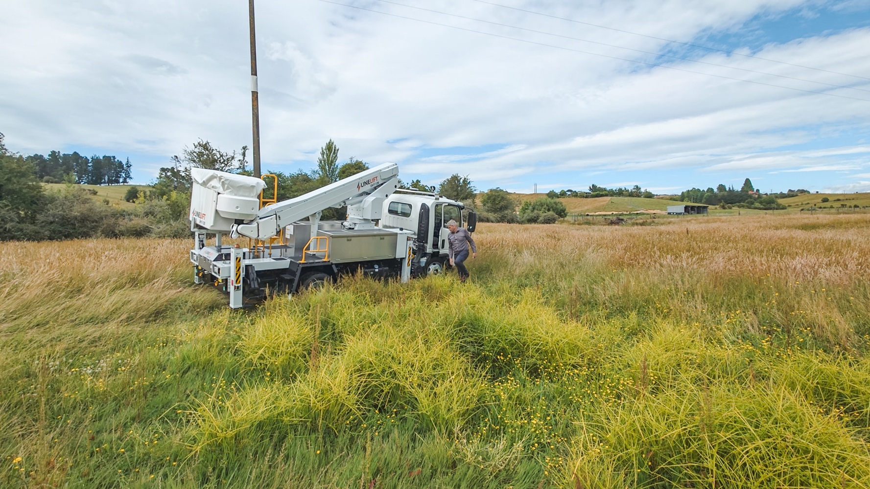 A specialist utility truck’s right rear outrigger is deployed on a grassy slope with a stabiliser pad and wedge to support it, with mudflaps on the wheels in the background with Waimea Group branding on them.
