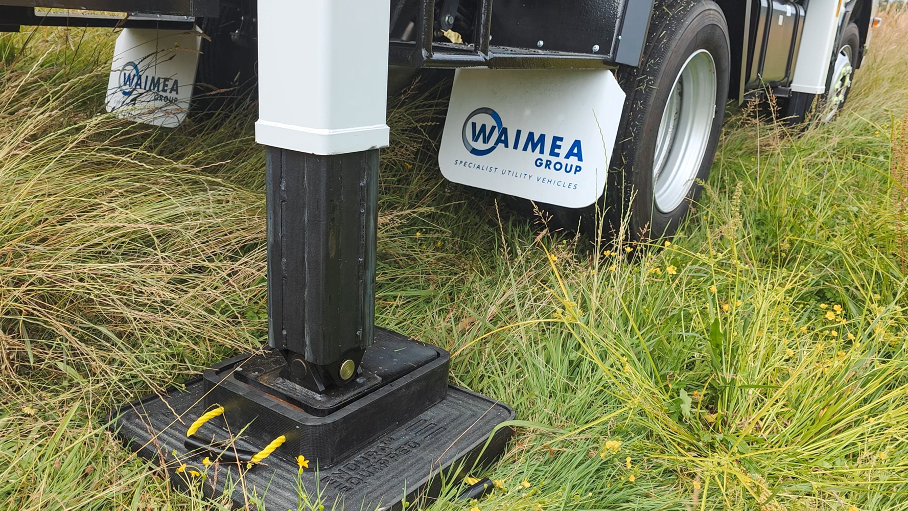 A specialist utility truck’s right rear outrigger is deployed on a grassy slope with a stabiliser pad and wedge to support it, with mudflaps on the wheels in the background with Waimea Group branding on them.