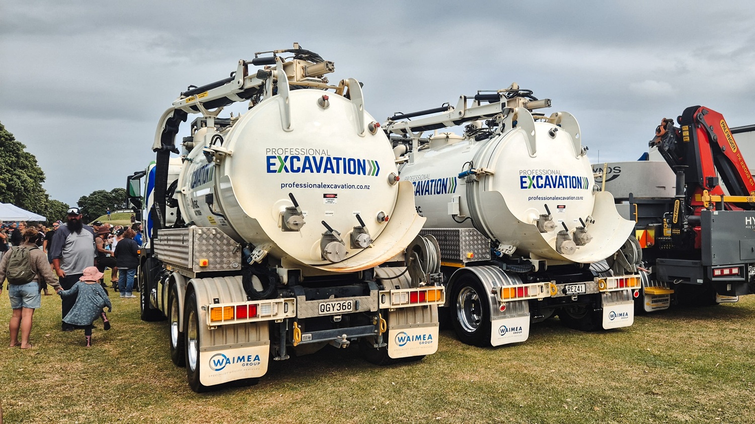 Two large white Allvac® hydrovac trucks with tanks and hose reels mounted on the back exiting the gate to a workyard. They are signwritten with Professional Excavation’s blue and green branding.