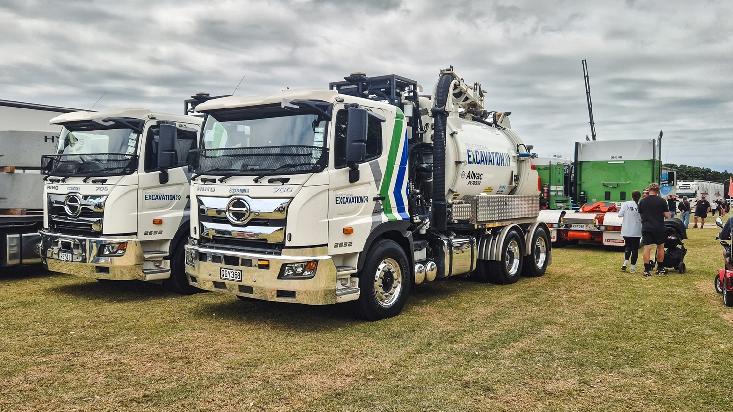 Two large white Allvac® hydrovac trucks with tanks and hose reels mounted on the back exiting the gate to a workyard. They are signwritten with Professional Excavation’s blue and green branding.