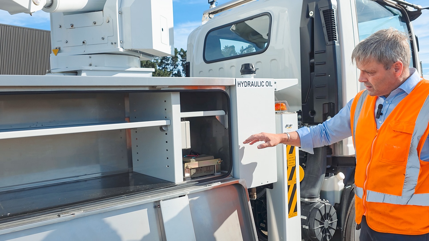 A man in a hi-vis vest stands in front of a white Linelift® bucket truck, which is parked in the background and has the boom and bucket extended into the foreground. The man is gesturing to the bucket.