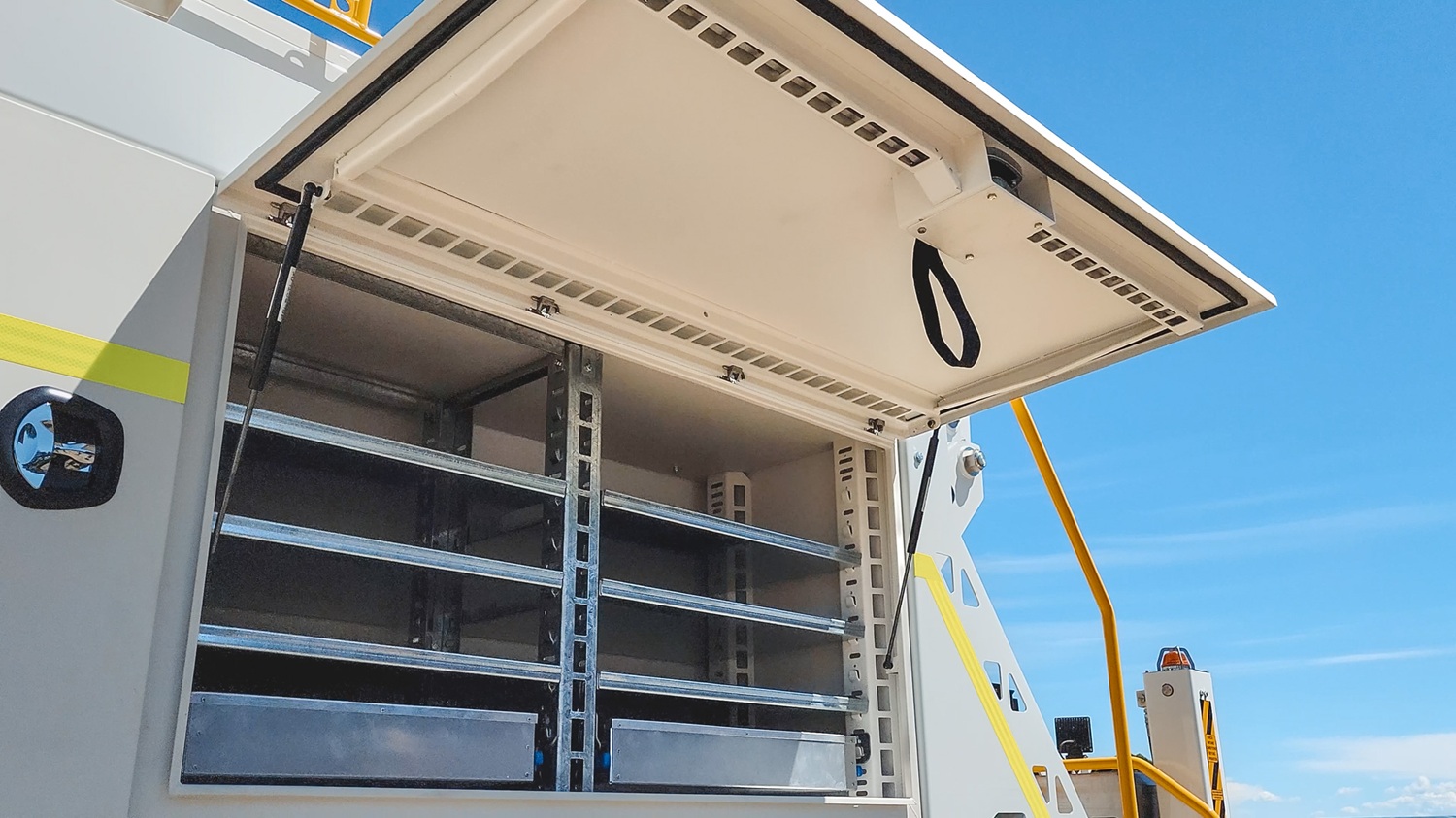 The side of a specialist utility EWP truck, angled towards the camera in front of an empty area with a blue sky behind it. The focus is on the open white toolboxes mounted on the side of the truck with shelving and room for equipment inside.