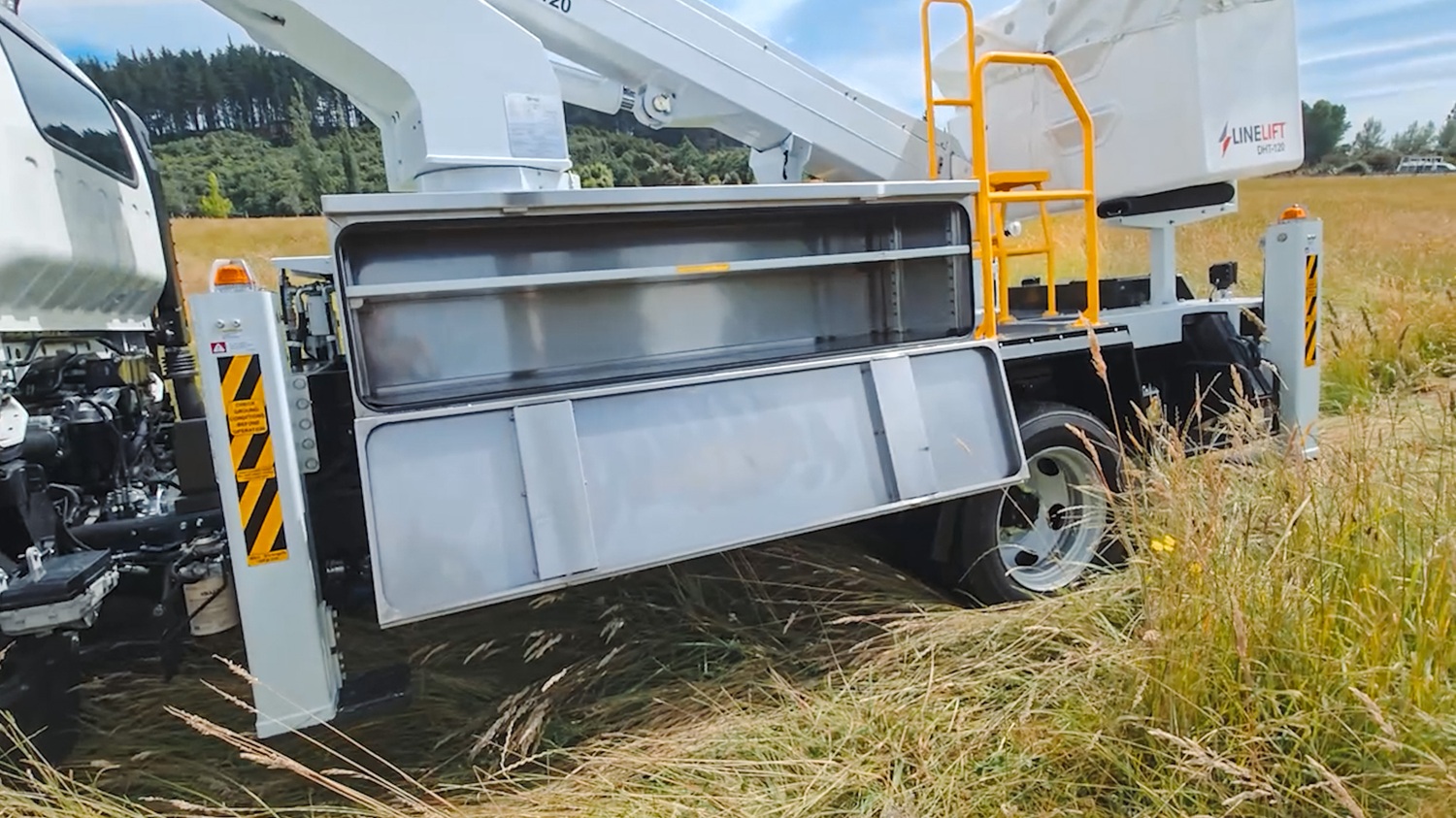 The side of a specialist utility EWP truck, angled towards the camera in front of an empty area with a blue sky behind it. The focus is on the open white toolboxes mounted on the side of the truck with shelving and room for equipment inside.