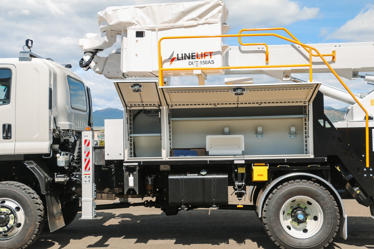 The side of a specialist utility EWP truck, angled towards the camera in front of an empty area with a blue sky behind it. The focus is on the open white toolboxes mounted on the side of the truck with shelving and room for equipment inside.