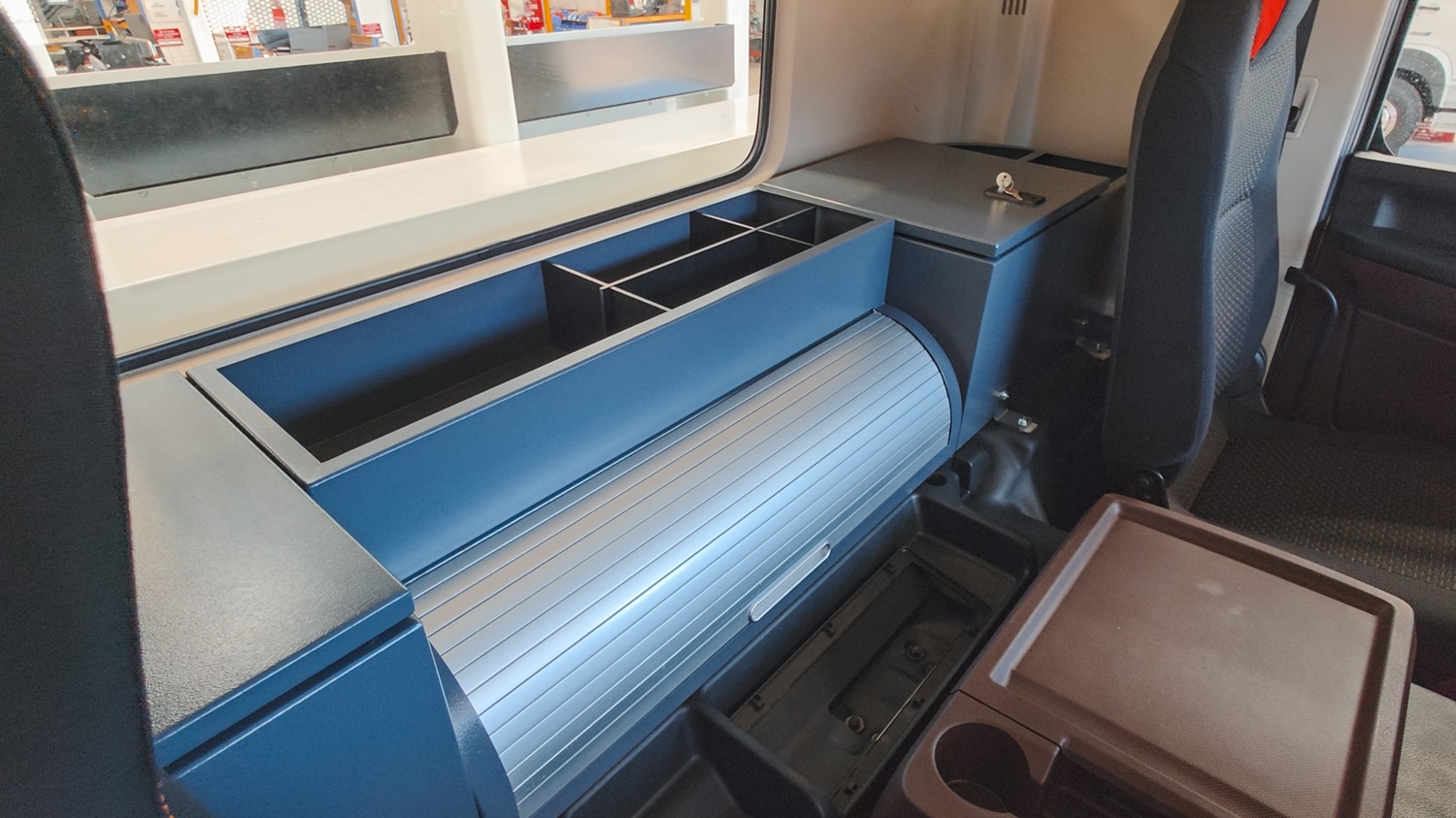 The side of a specialist utility EWP truck, angled towards the camera in front of an empty area with a blue sky behind it. The focus is on the open white toolboxes mounted on the side of the truck with shelving and room for equipment inside.