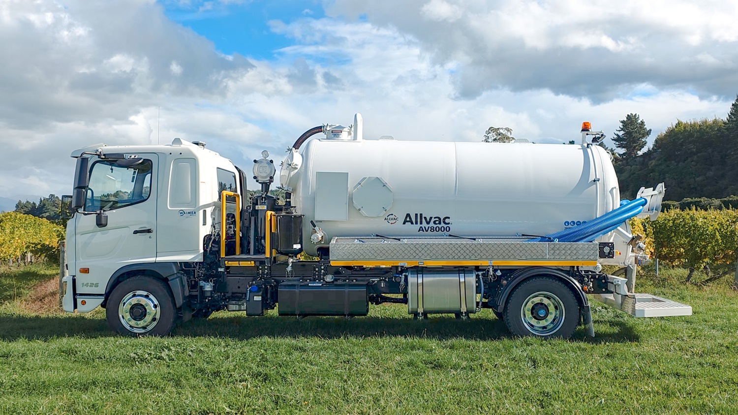 A large white vacuum tanker truck with Allvac® branding on the side, blue hoses in side hose trays, and an access platform at the front of the tank, parked on the grass in front of a vineyard.