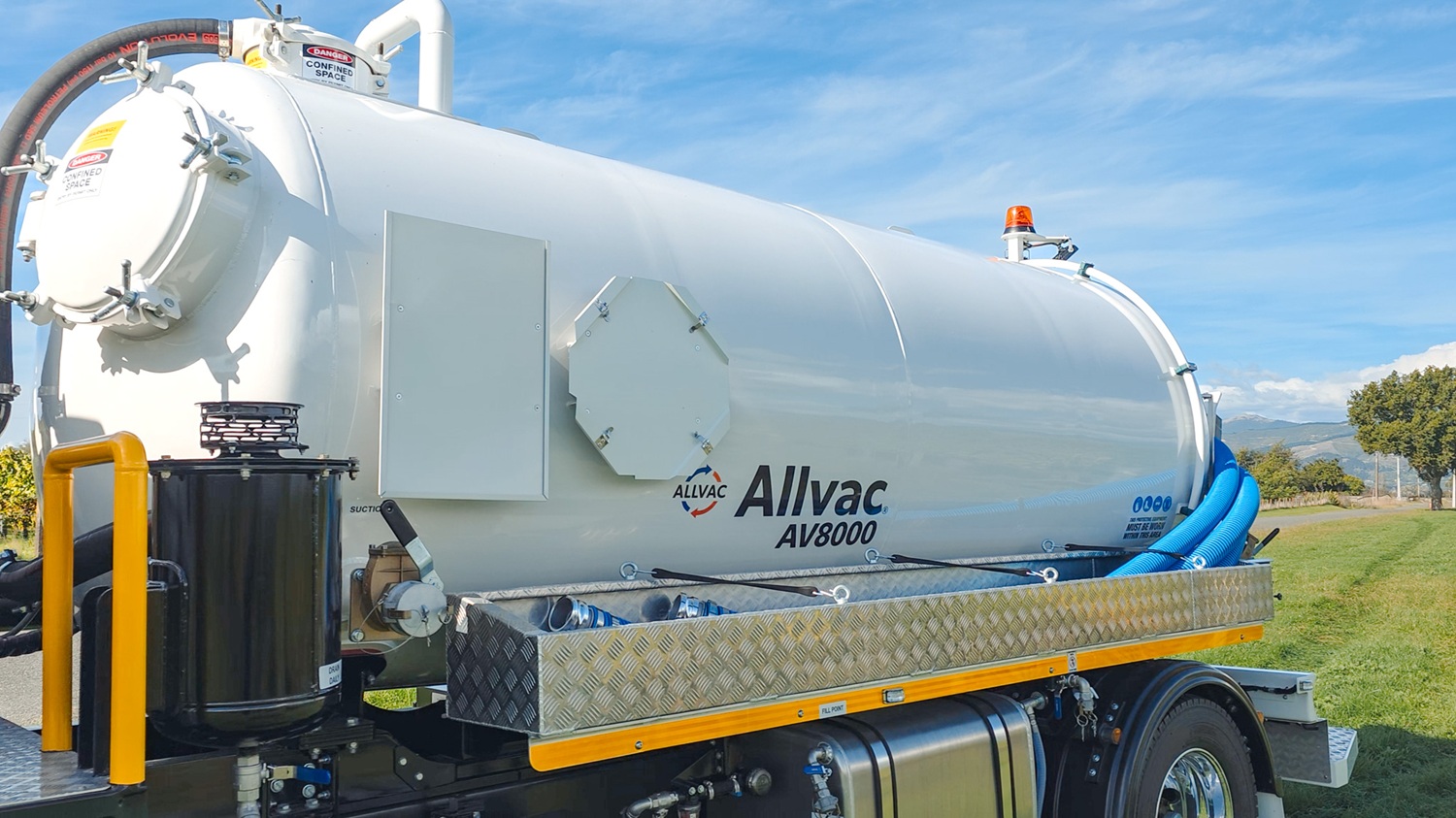 A large white vacuum tanker truck with Allvac® branding on the side, blue hoses in side hose trays, and an access platform at the front of the tank, parked on the grass in front of a vineyard.