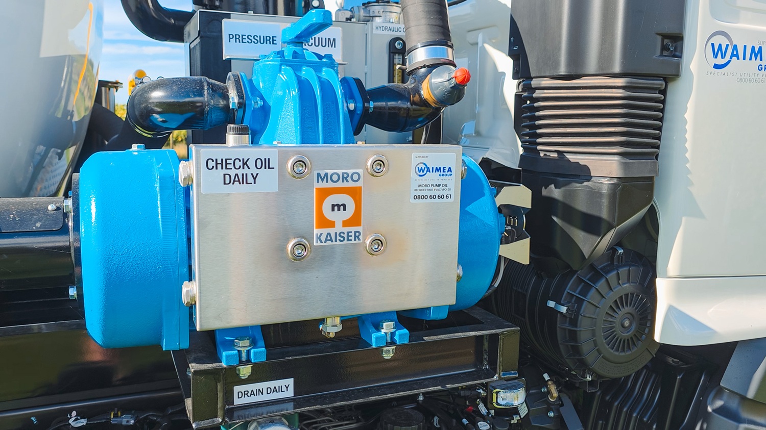 A large white vacuum tanker truck with Allvac® branding on the side, blue hoses in side hose trays, and an access platform at the front of the tank, parked on the grass in front of a vineyard.