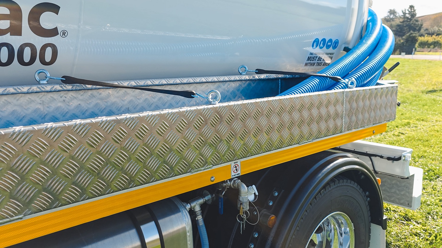 A large white vacuum tanker truck with Allvac® branding on the side, blue hoses in side hose trays, and an access platform at the front of the tank, parked on the grass in front of a vineyard.