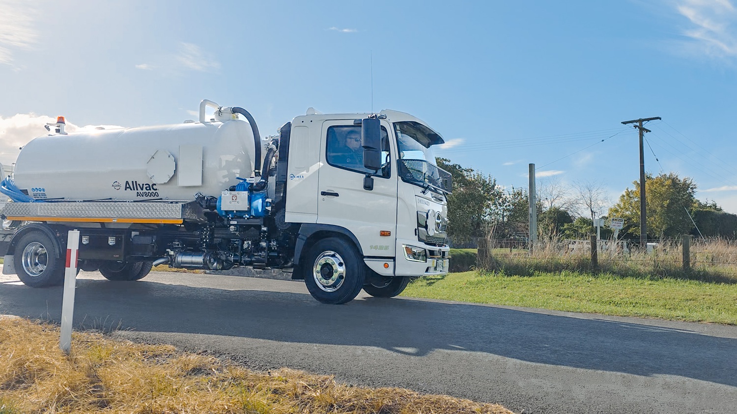 A large white vacuum tanker truck with Allvac® branding on the side, blue hoses in side hose trays, and an access platform at the front of the tank, parked on the grass in front of a vineyard.