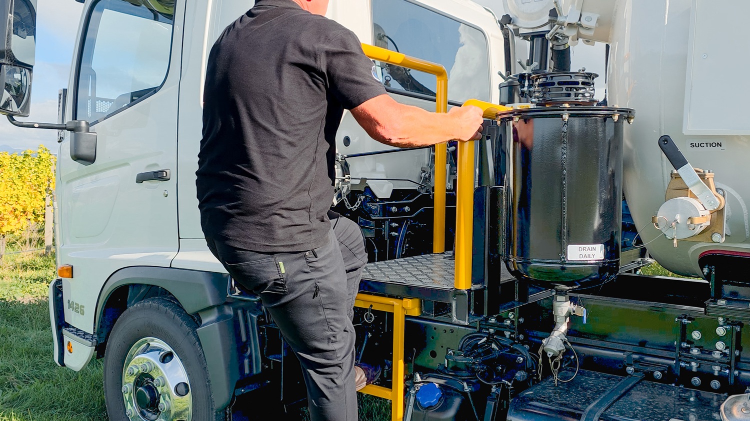 A large white vacuum tanker truck with Allvac® branding on the side, blue hoses in side hose trays, and an access platform at the front of the tank, parked on the grass in front of a vineyard.