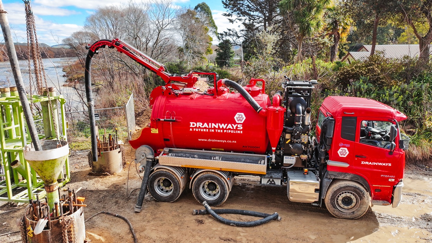 A large red hydrovac truck is parked on a riverbank with other excavation equipment, excavating holes for bridge foundations.