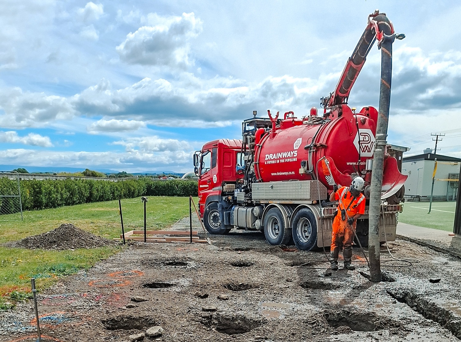 A large red hydrovac truck is parked on a riverbank with other excavation equipment, excavating holes for bridge foundations.