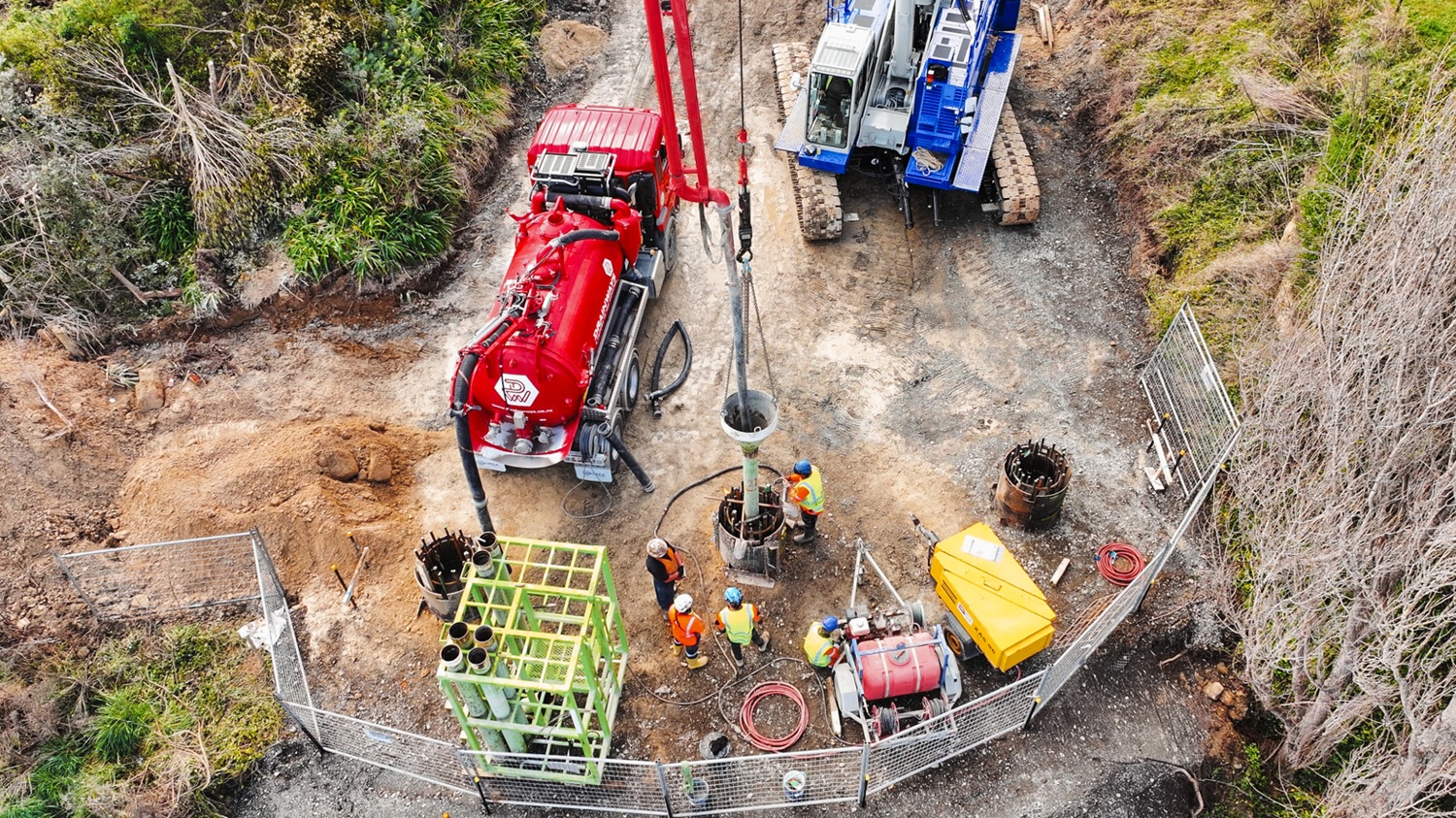 A large red hydrovac truck is parked on a riverbank with other excavation equipment, excavating holes for bridge foundations.