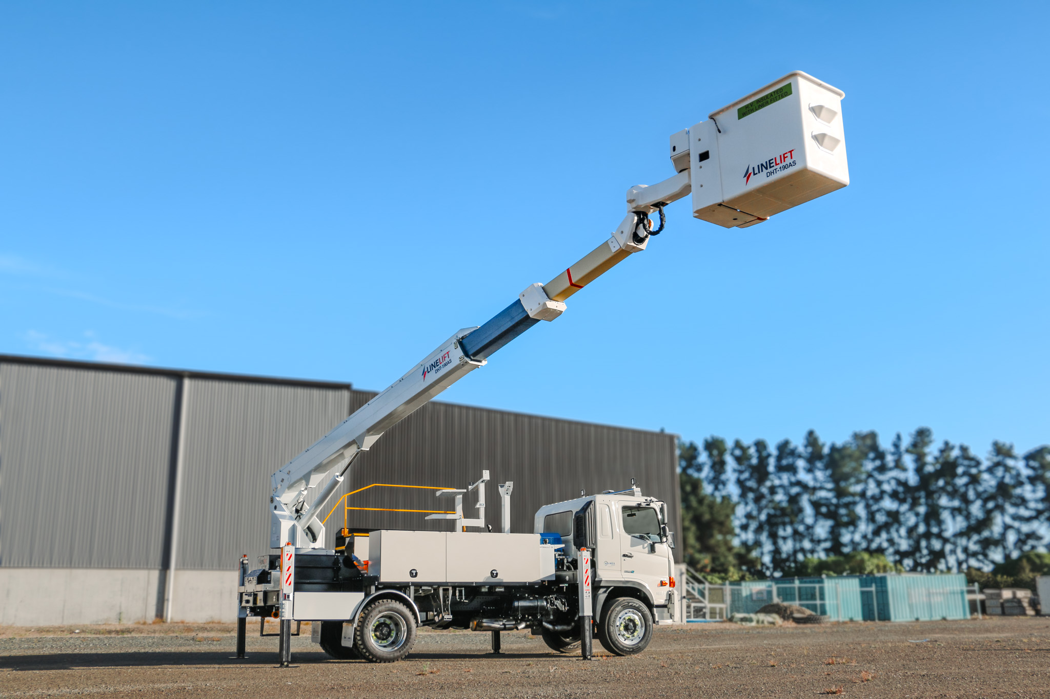 A white EWP truck parked in an empty lot, with a powerful Hino chassis, traction tyres, large sidemounted toolboxes, and an insulated bucket boom mounted on the back with Linelift DHT190AS branding, accessed by steps from the back with a railing.