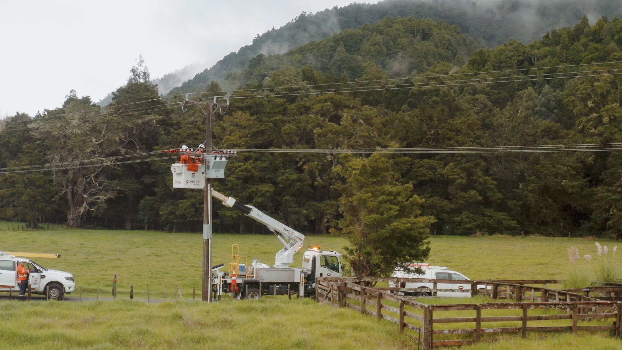 An EWP with a telescopic boom working on power lines in a field next to the treeline, with support utes around it.