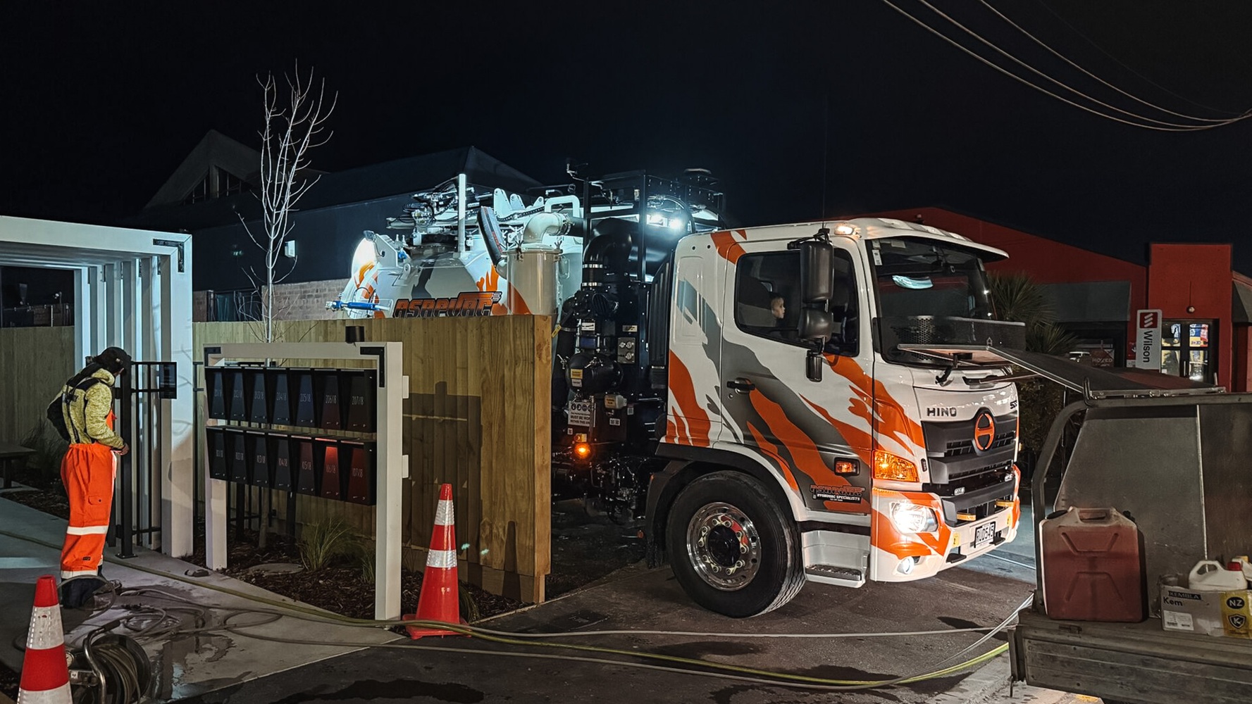 A white hydro excavation truck with custom signwriting in a parking lot with a dark cloudy sky behind it, showing the Allvac tank, overhead boom, and toolbox setup mounted on a Hino cab-chassis lit up by truck lights.