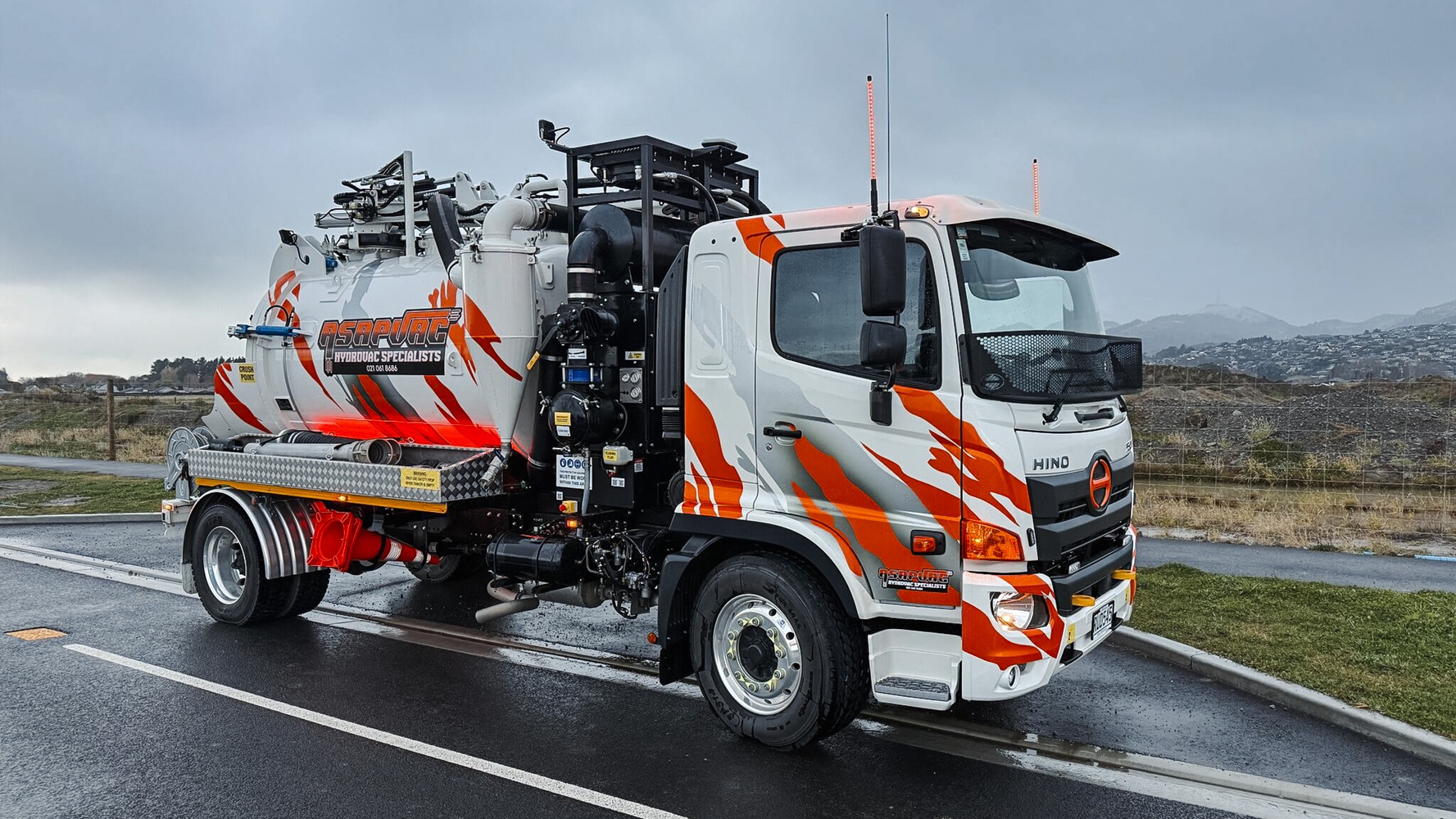 A white hydro excavation truck with custom signwriting in a parking lot with a dark cloudy sky behind it, showing the Allvac tank, overhead boom, and toolbox setup mounted on a Hino cab-chassis lit up by truck lights.