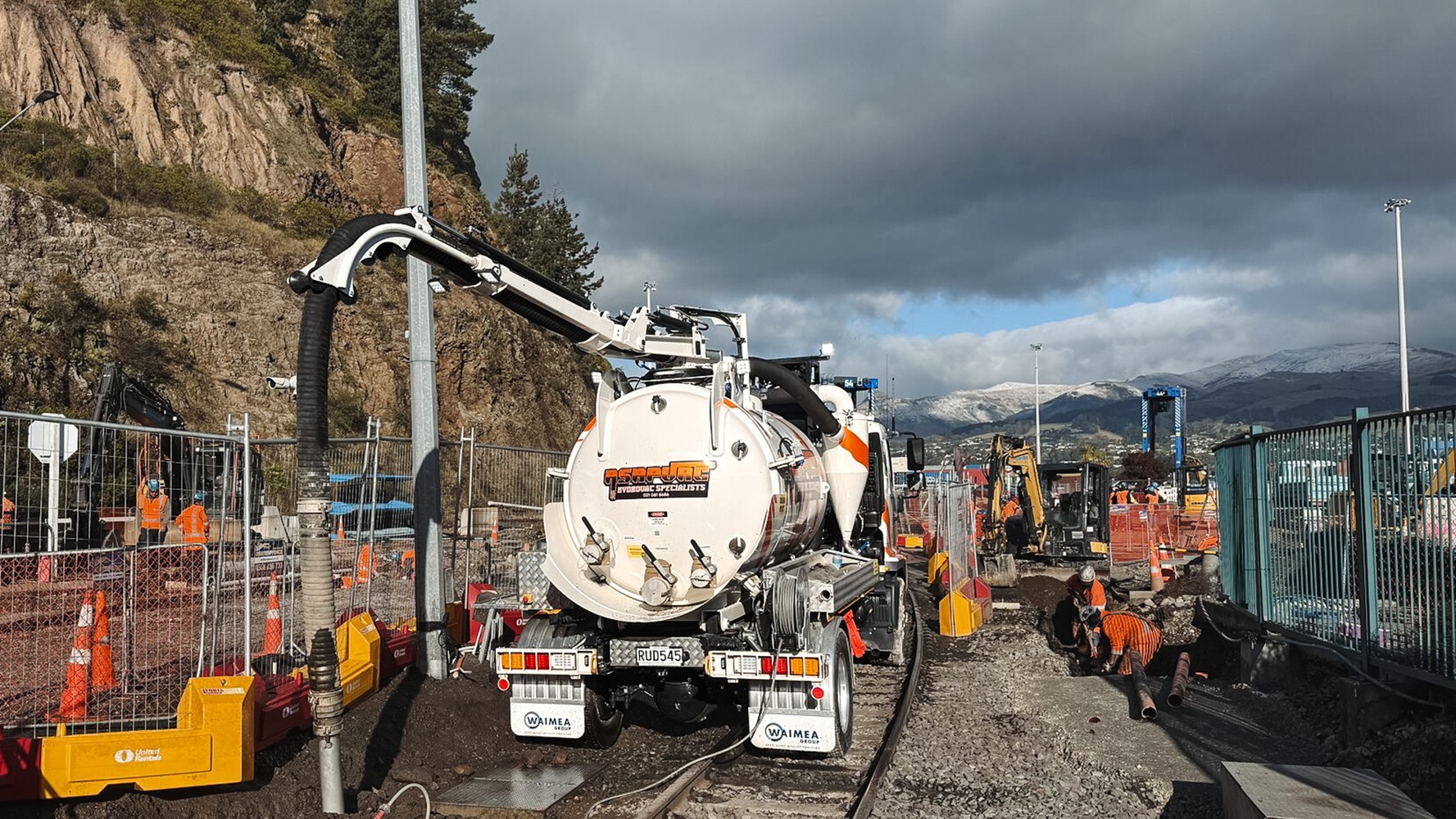 A white hydro excavation truck with custom signwriting in a parking lot with a dark cloudy sky behind it, showing the Allvac tank, overhead boom, and toolbox setup mounted on a Hino cab-chassis lit up by truck lights.