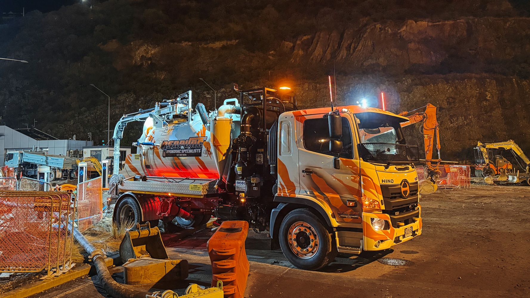 A white hydro excavation truck with custom signwriting in a parking lot with a dark cloudy sky behind it, showing the Allvac tank, overhead boom, and toolbox setup mounted on a Hino cab-chassis lit up by truck lights.