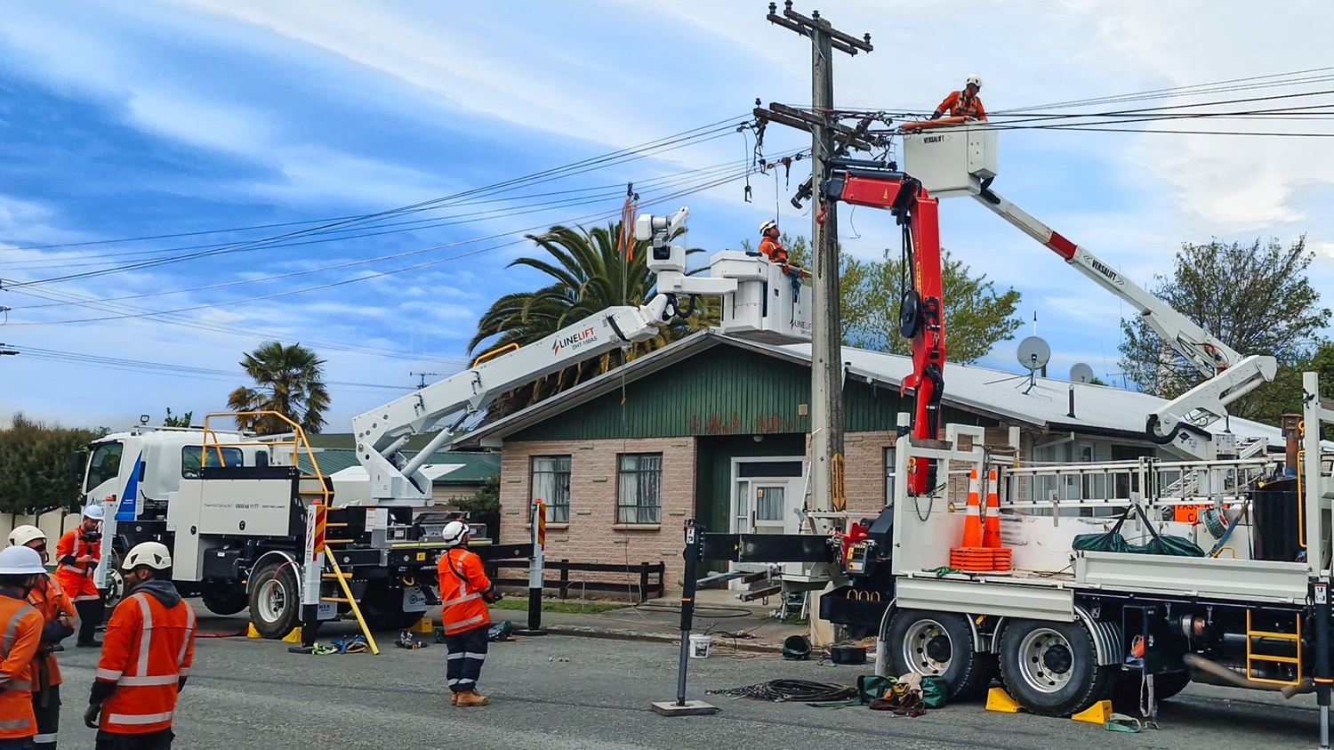 A white Isuzu 4x4 bucket truck parked on the side of the road in a suburban area, with its outriggers down and the insulated boom extended down to the ground next to a power pole.