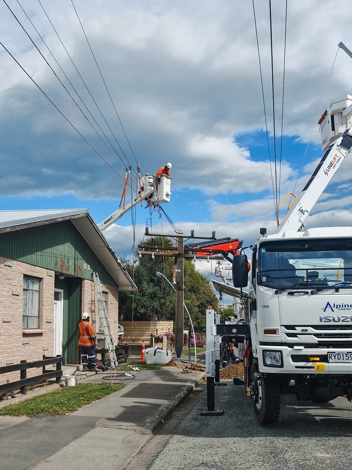 A white Isuzu 4x4 bucket truck parked on the side of the road in a suburban area, with its outriggers down and the insulated boom extended down to the ground next to a power pole.