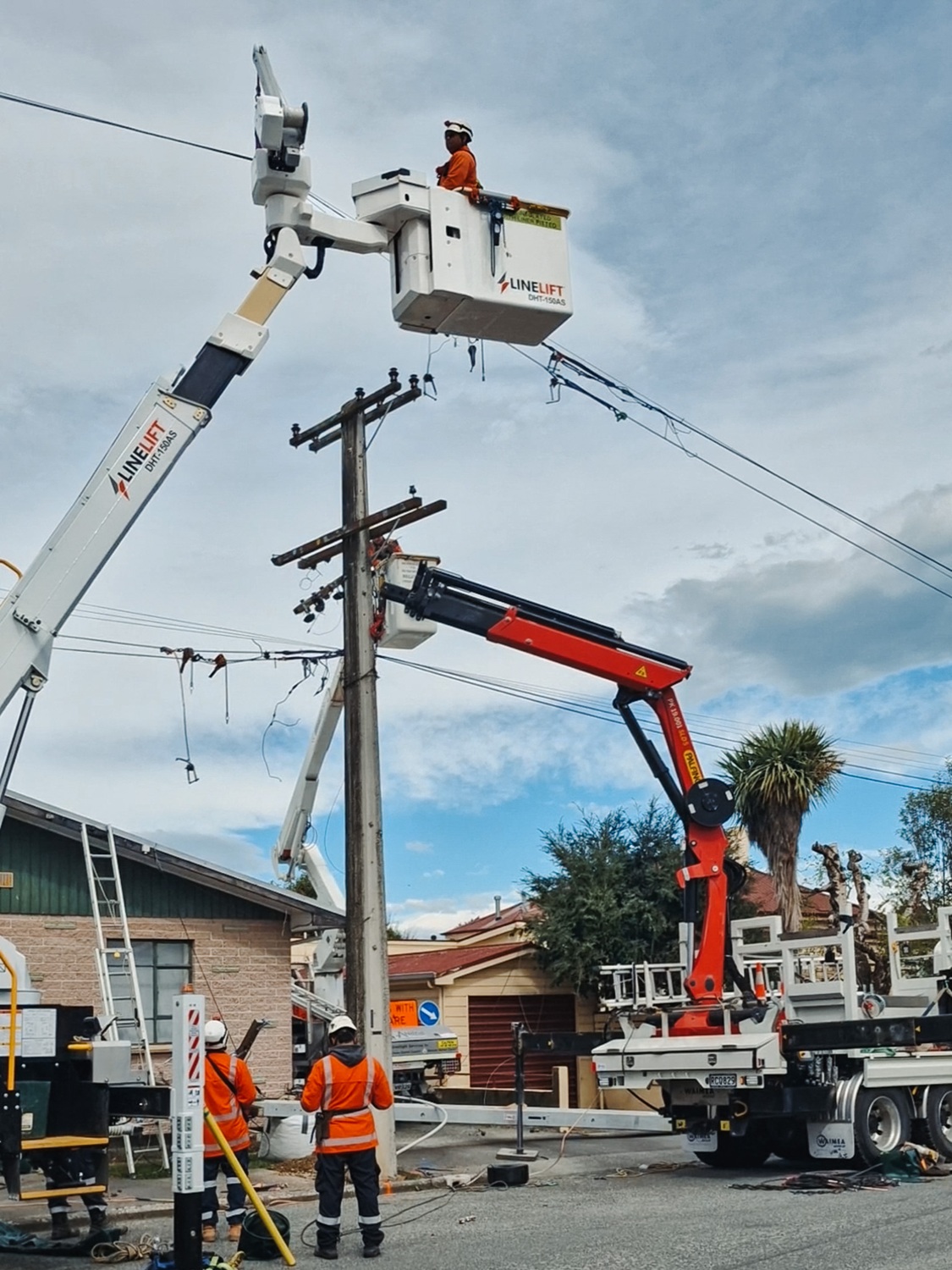 A white Isuzu 4x4 bucket truck parked on the side of the road in a suburban area, with its outriggers down and the insulated boom extended down to the ground next to a power pole.