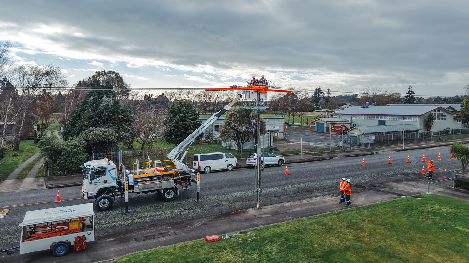 An aerial view of three white Scanpower trucks parked along a rural road beside a green hillside, with two insulated bucket booms raised to a new pole structure while line workers in orange hi-vis work from the buckets.