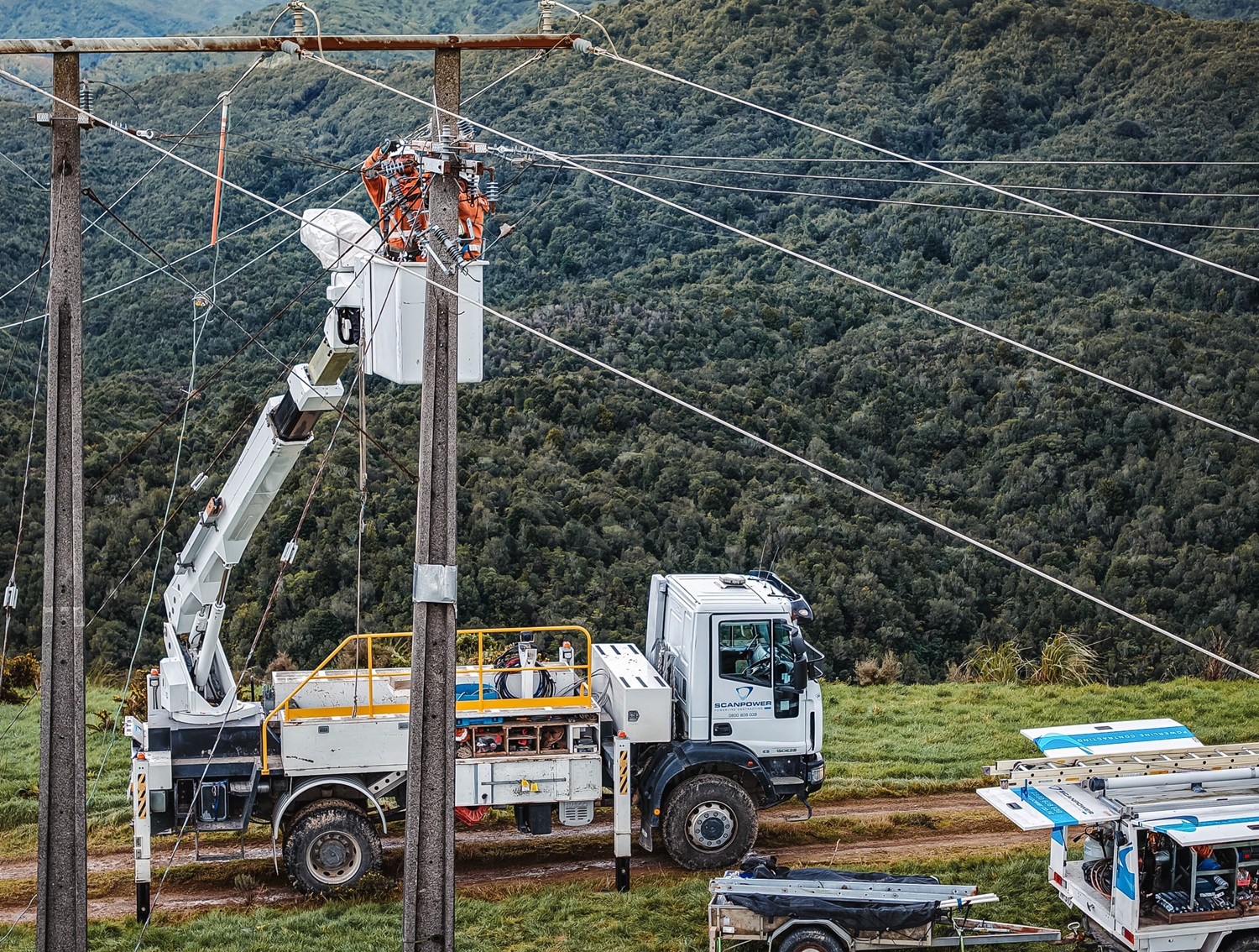 An aerial view of three white Scanpower trucks parked along a rural road beside a green hillside, with two insulated bucket booms raised to a new pole structure while line workers in orange hi-vis work from the buckets.